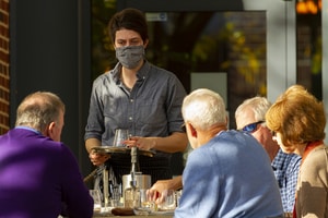 A young waitress wearing a face mask due to COVID-19 is serving at an outdoor table where elderly customers who are not wearing mask are sitting close to each other.